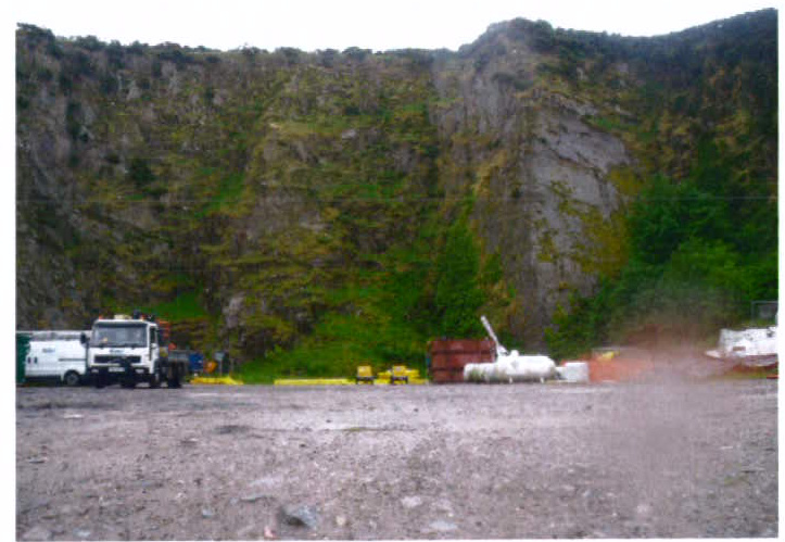 A photograph showing a gravel site with industrial vehicles and equipment in front of a steep, rocky cliff face covered in vegetation.
