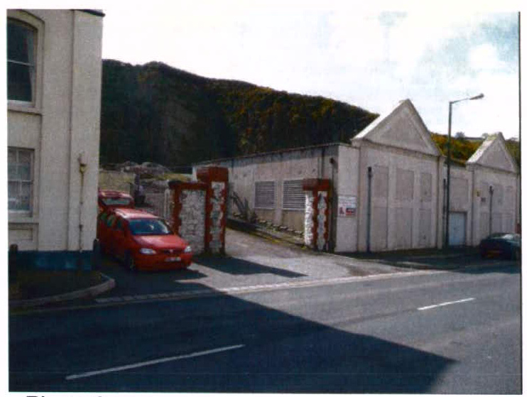 A street-level photograph showing a white building with gabled ends, likely garages, next to a larger white building with a red car parked nearby.