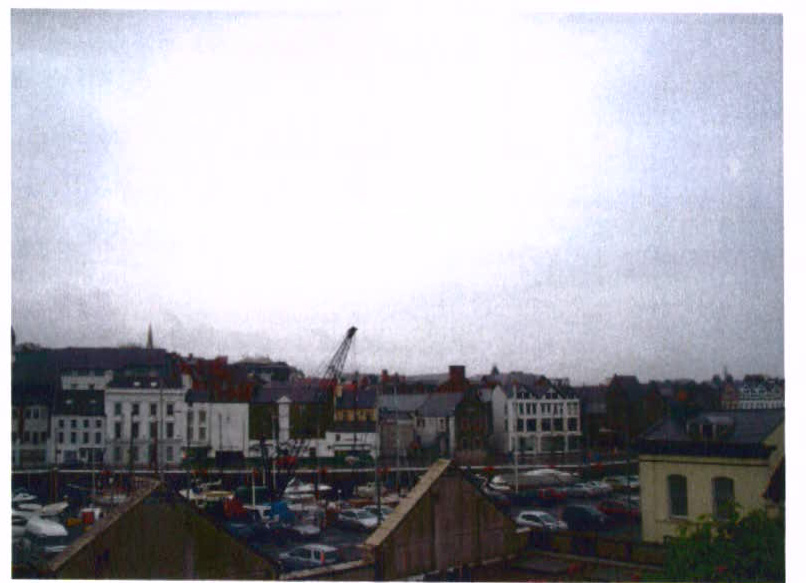 A grainy photograph showing a waterfront or harbor area with various buildings, a crane, and parked vehicles or boats in the foreground.