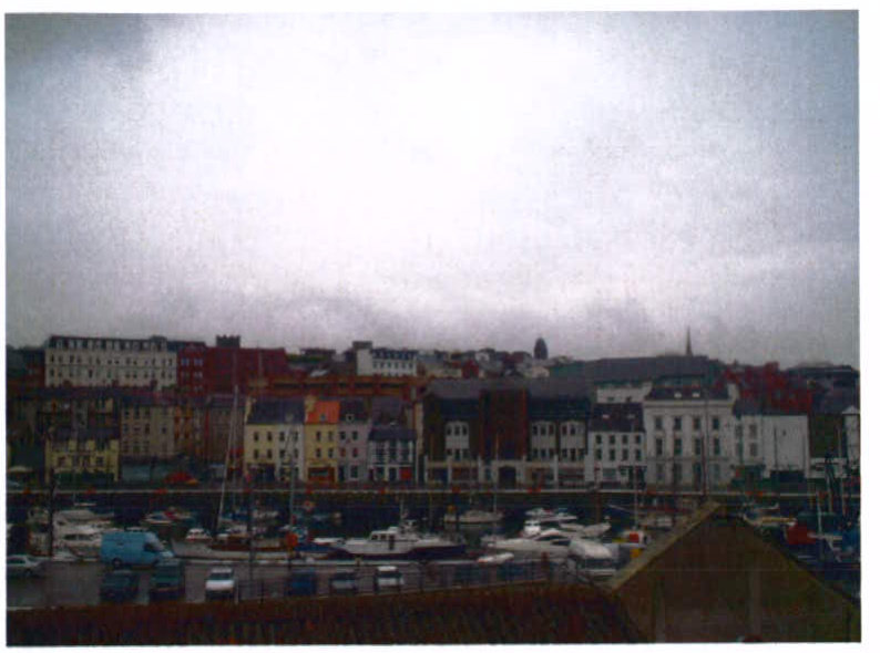 A grainy photograph showing a harbor or marina with boats in the foreground and a row of multi-story buildings along the waterfront in the background under a cloudy sky.