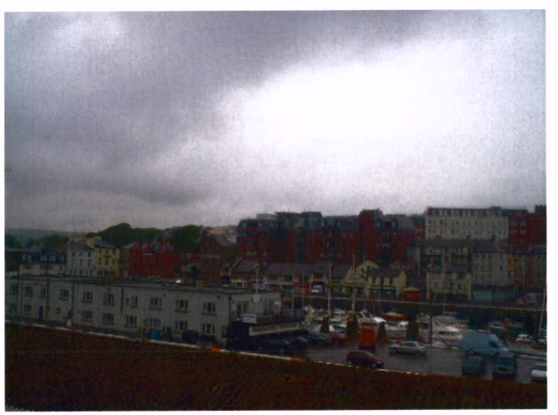 A photograph showing an elevated view of a town with multi-story commercial buildings, a parking area, and an overcast sky.