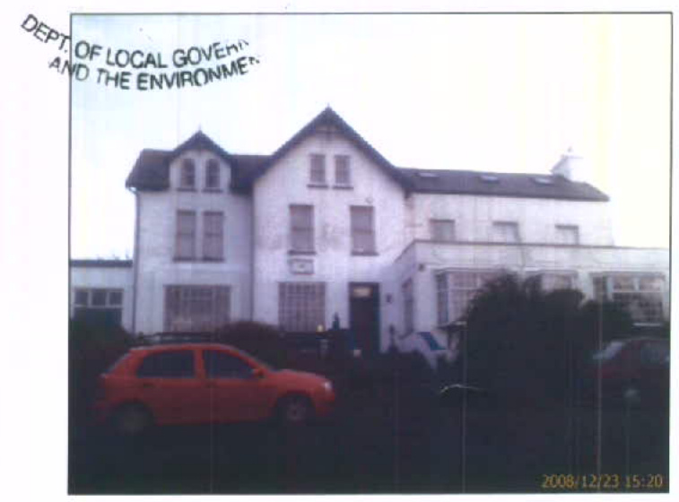 A photograph showing the exterior of a large white detached building, likely a former hotel, with a red car parked in the foreground.