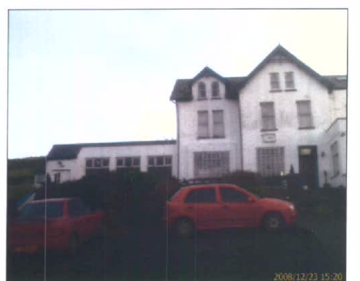 A grainy exterior photograph of a white, multi-story building complex, likely a former hotel, with red cars parked in the foreground.