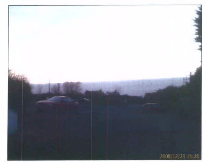A grainy photograph showing a car parked on a driveway or road with a view of the sea in the background.