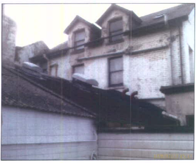 A grainy photograph showing the upper facade and roofline of a white building with dormer windows, viewed from behind a lower dark roof structure.