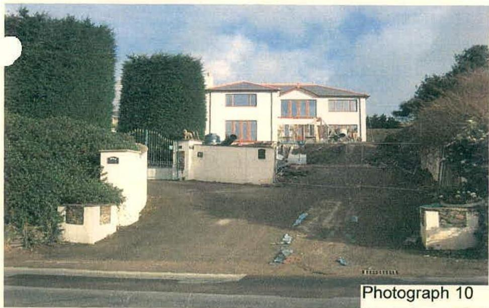 A photograph showing a white, two-story detached house with a driveway, boundary walls, and surrounding vegetation.