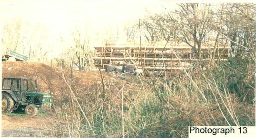 A photograph showing a rural construction site with a tractor in the foreground and a timber-framed building structure under construction in the background.