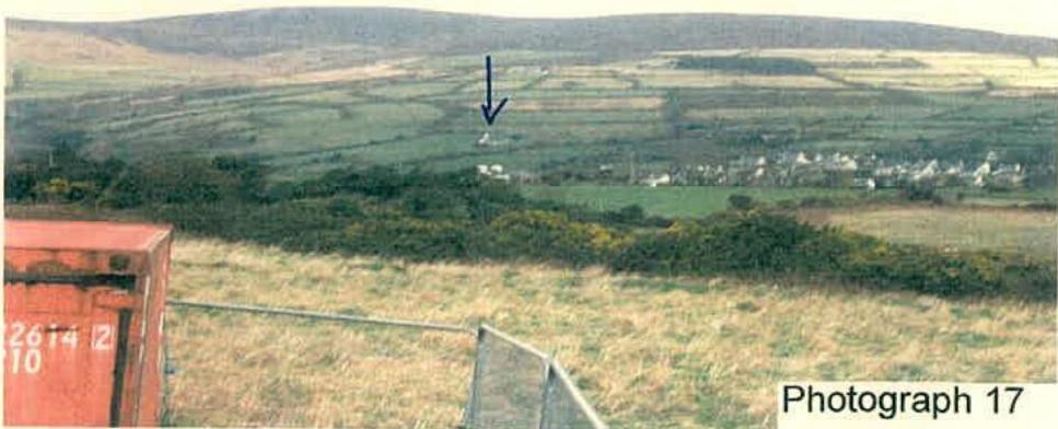 A landscape photograph showing a rural valley with a black arrow pointing to a distant white building, likely indicating the site location.
