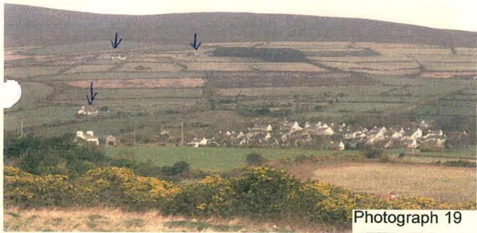 A landscape photograph showing a rural valley with fields and a settlement, featuring blue arrows pointing to specific buildings on the hillside.