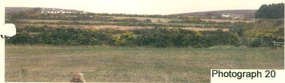 A wide-angle photograph showing the rural surroundings of the site, featuring grassy fields, dense hedgerows, and distant buildings on a hillside.