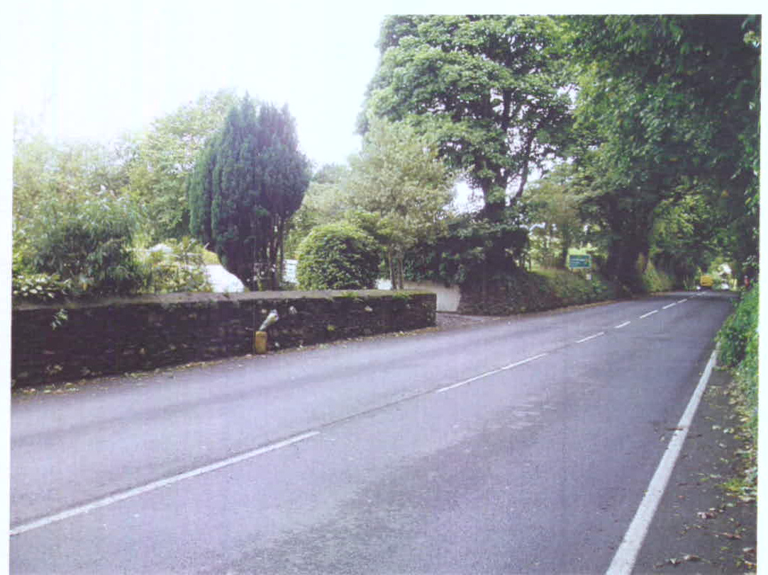 A photograph showing a paved road with a stone wall and dense vegetation on the left side, depicting a rural setting with a potential driveway entrance.