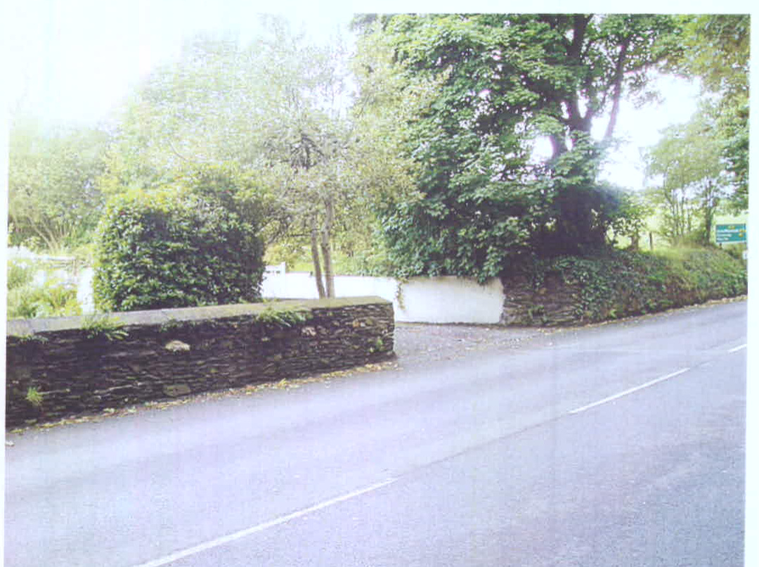 A photograph showing a roadside view featuring a low stone boundary wall and a driveway entrance surrounded by trees and vegetation.