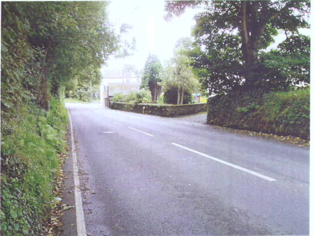 A photograph showing a paved rural road curving to the left, bordered by a stone wall and dense vegetation, likely depicting the existing access point.