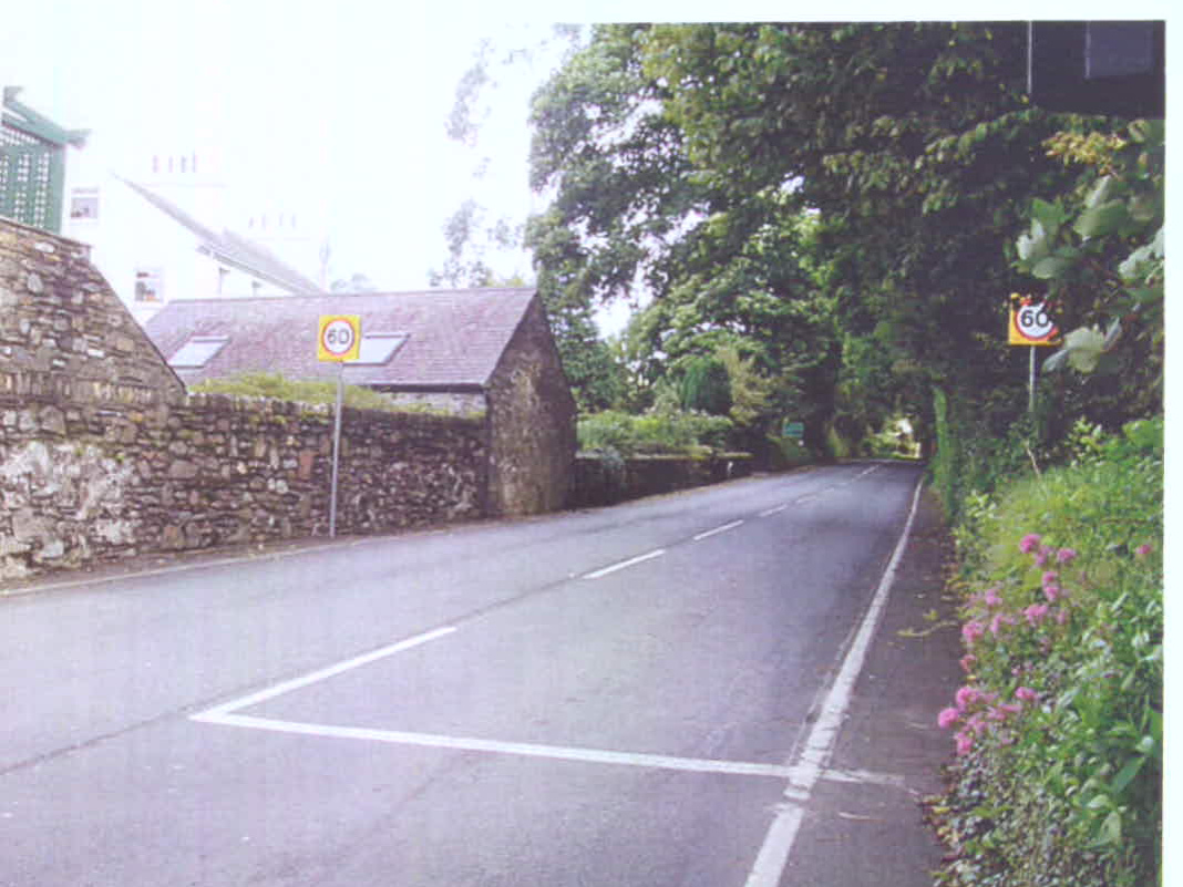 A photograph showing a rural road scene with a stone wall and a stone outbuilding on the left side, featuring a 60mph speed limit sign.