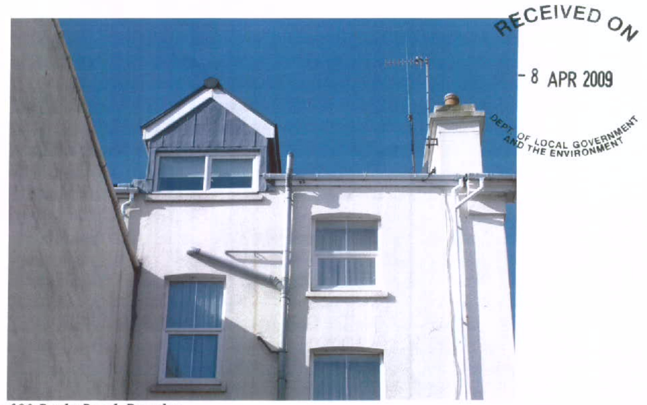 A photograph showing the upper facade of a white residential building featuring a dormer window on the roof against a blue sky.