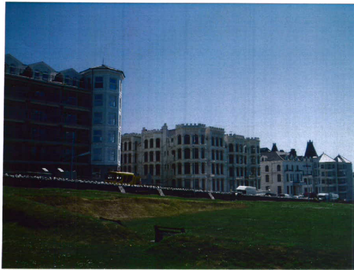 A photograph showing a row of large, multi-story buildings along a waterfront with a grassy slope in the foreground.