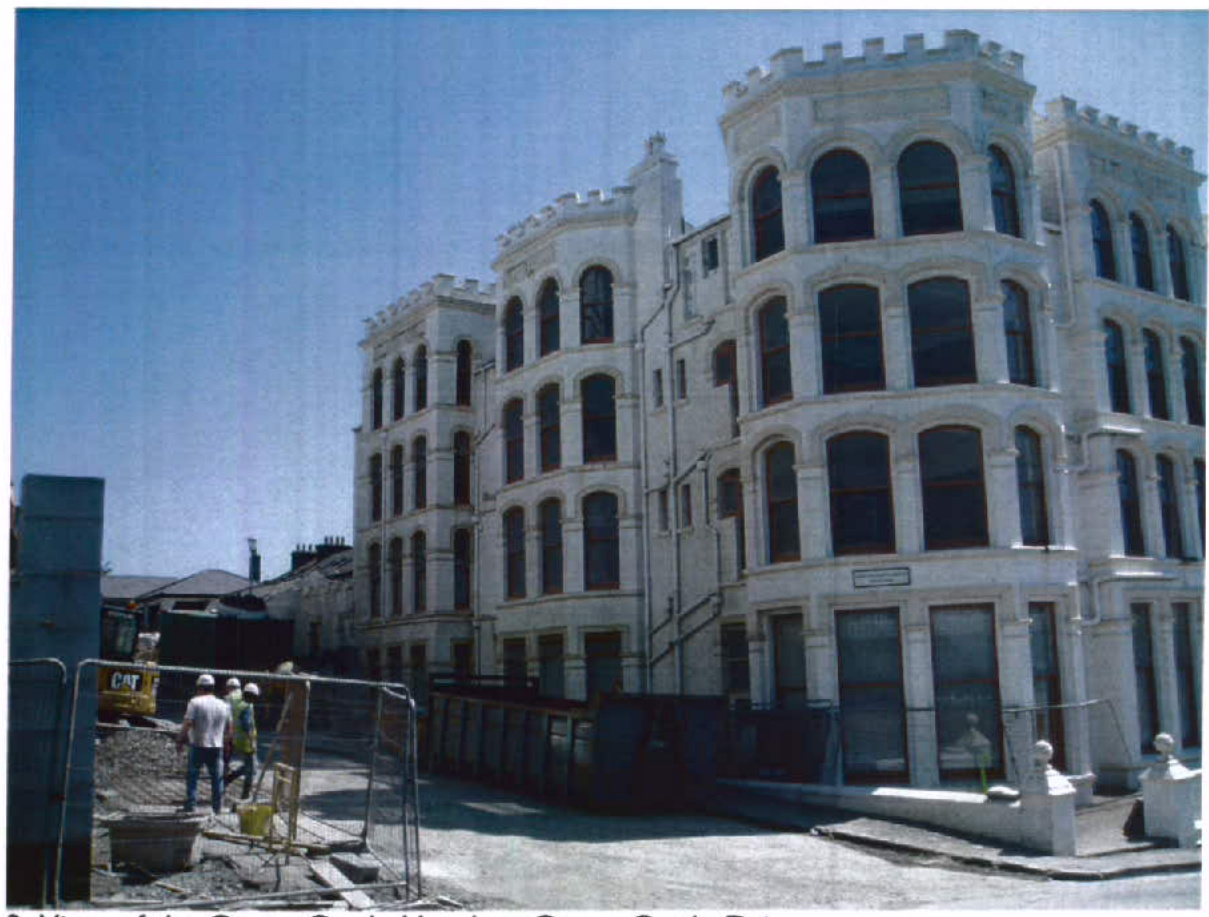 A photograph showing a large, white, multi-story building with crenellated rooflines under construction, featuring workers and machinery in the foreground.