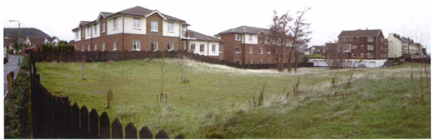 A panoramic photograph showing a grassy, undeveloped plot of land in the foreground bordered by a wooden fence, with existing residential buildings visible in the background.