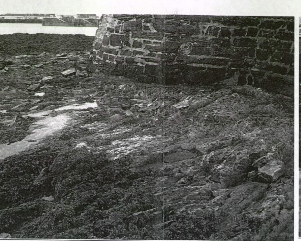 A black and white photograph showing a rocky, uneven coastal ground surface with a large stone wall structure in the background.