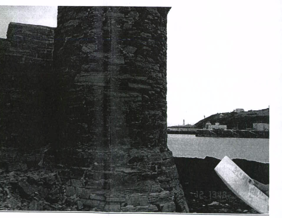 A grainy black and white photograph showing a close-up of a stone tower or wall section with a harbor and hillside in the background.