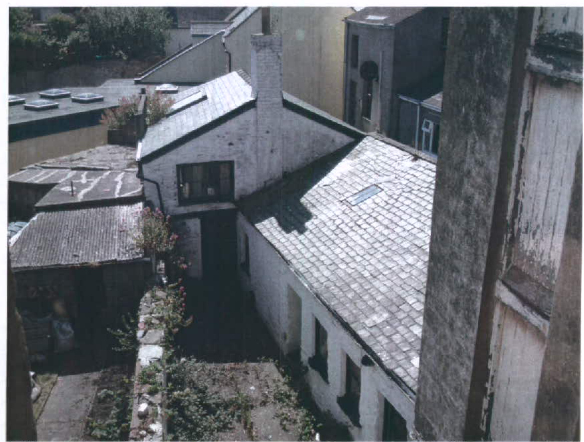 A high-angle photograph showing a white-washed building with a slate roof and skylights, adjacent to a lower outbuilding with a corrugated roof and a garden area.