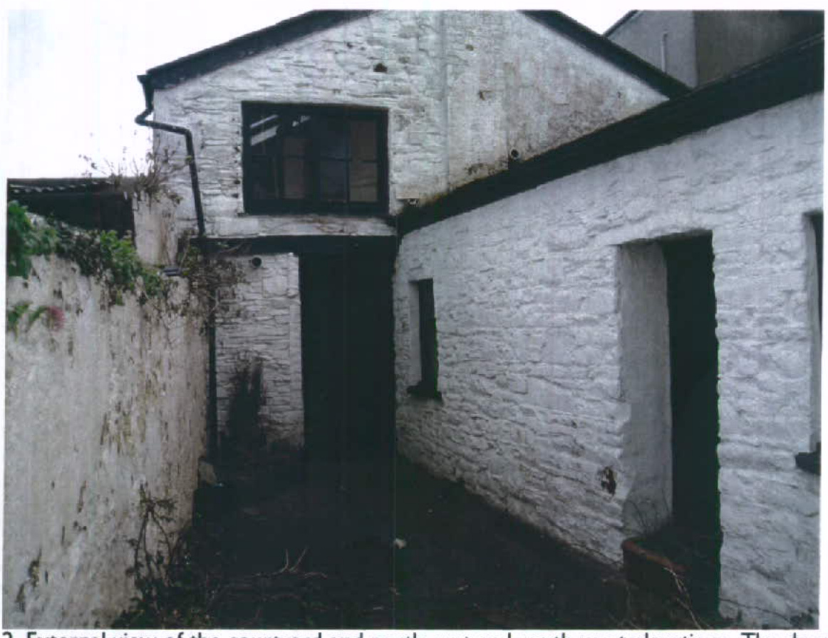 External photograph of a white-washed stone outbuilding showing a courtyard area and dark window and door openings.