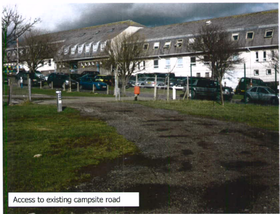 A photograph showing a gravel track leading to a parking area with a long white building in the background, captioned 'Access to existing campsite road'.