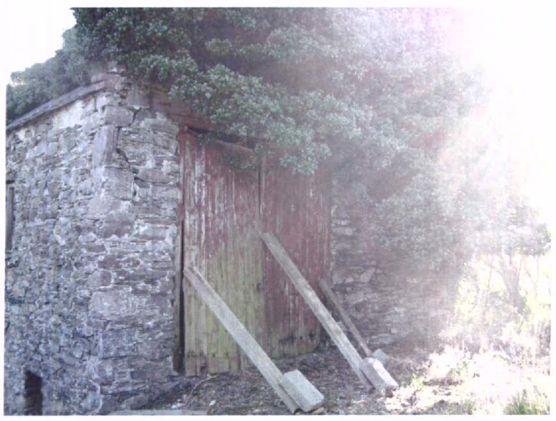 A photograph showing the exterior of an old stone building, likely a mill or barn, with large wooden doors and vegetation growing on the roof.