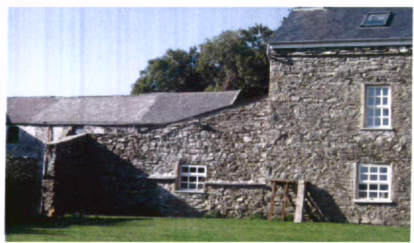 A photograph showing the exterior of a stone building with a slate roof and white-framed windows, situated in a rural setting with grass and trees.