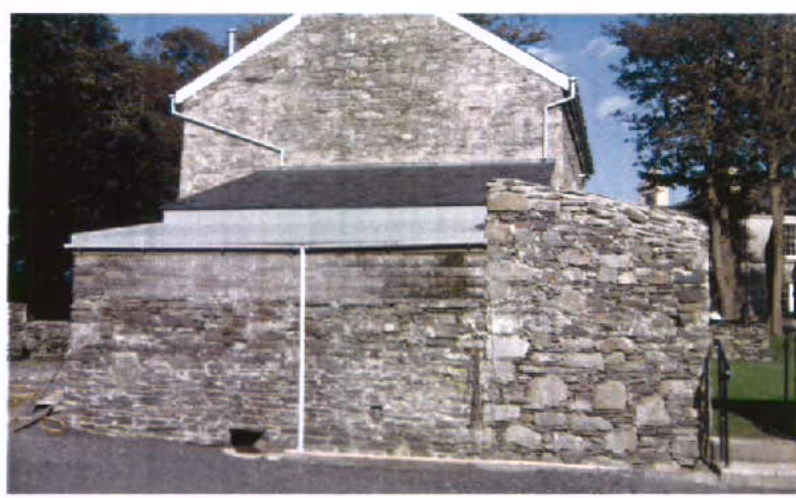 A photograph showing a stone building with a lower single-story extension featuring a slate roof and white guttering, situated next to a rough stone boundary wall.