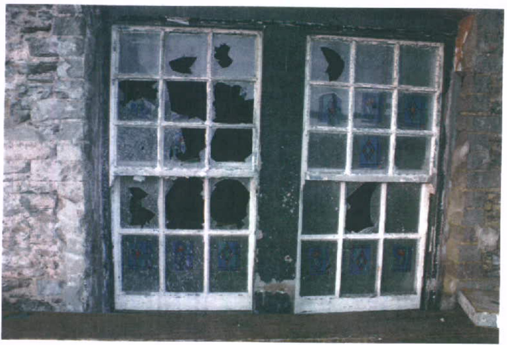 A photograph showing two damaged, multi-paned sash windows set in a rough stone wall, likely documenting the existing condition before replacement.