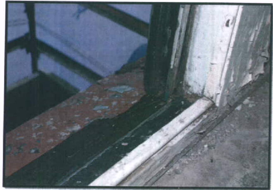 A close-up photograph showing the deteriorating condition of an existing wooden window frame and sill, with scaffolding visible in the background.
