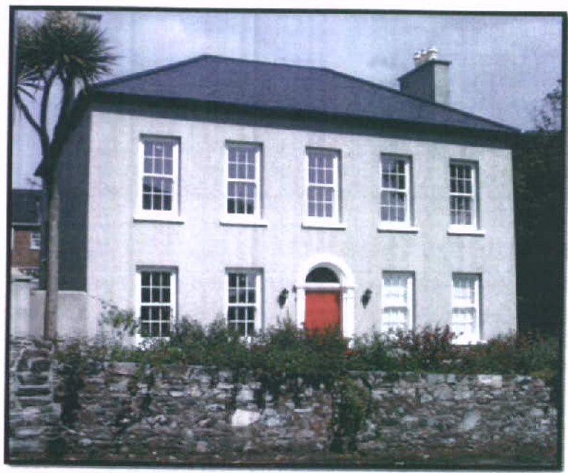 A photograph showing the front elevation of a two-story white house with sash windows and a red door, bordered by a stone wall and vegetation.