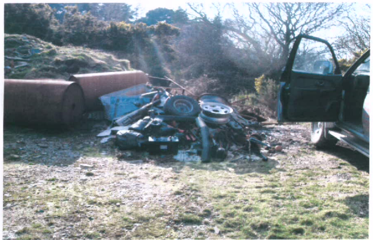 A photograph showing a pile of scrap metal, car parts, and large rusty cylinders in a grassy outdoor area.