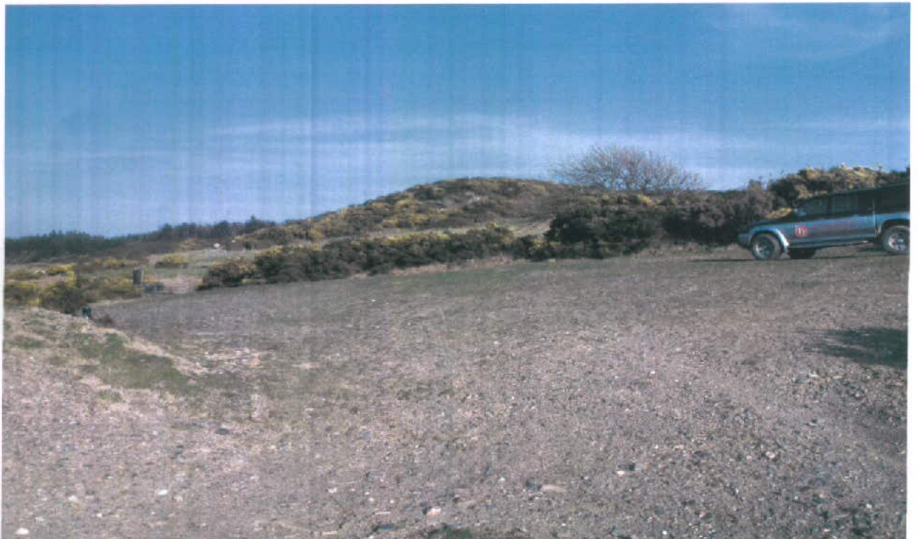 A photograph showing a gravelly open field with gorse bushes and a vehicle parked on the right side under a blue sky.