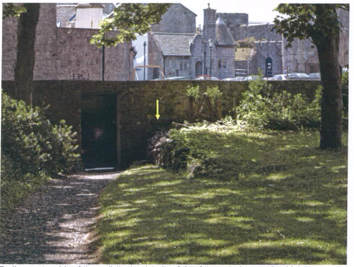 A photograph showing a stone boundary wall with a dark opening and a gravel path, with a yellow arrow pointing to a section of the wall near vegetation.