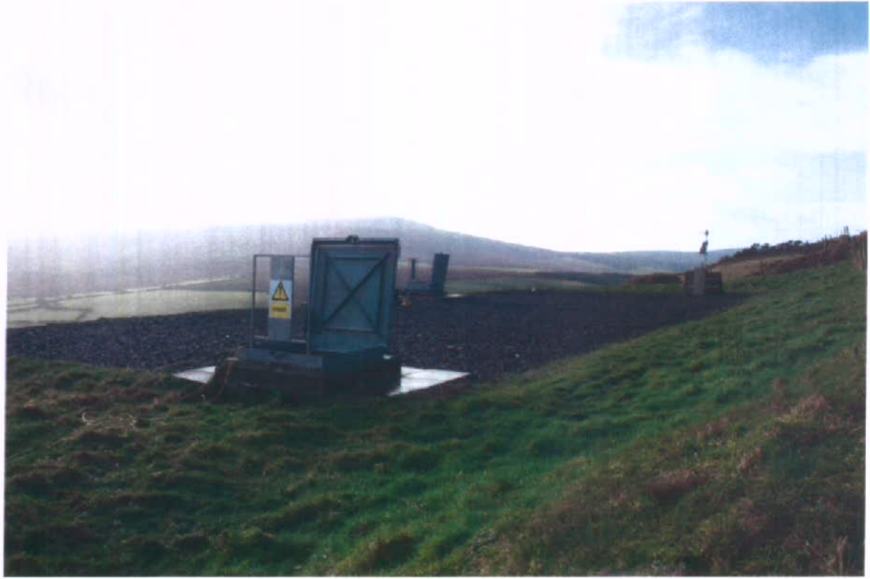 A photograph showing a grassy embankment with a concrete access structure and metal gate, likely part of a service reservoir, set against a rural backdrop.
