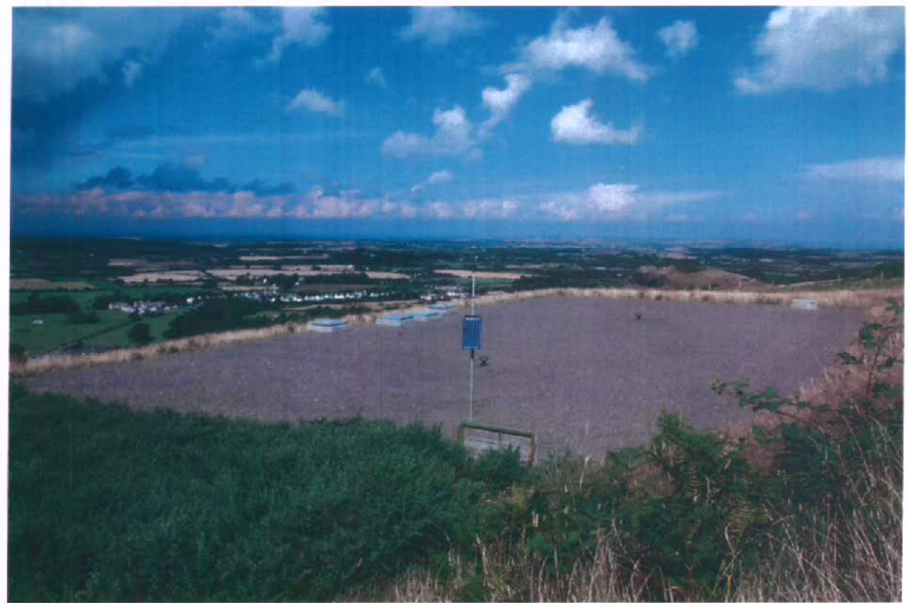 A wide-angle photograph showing a large, flat, gravel-covered service reservoir site in a rural setting with a village and sea visible in the background.