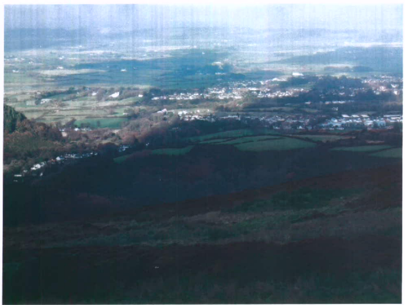 A high-angle photograph taken from a hilltop looking down over a rural valley with fields and a distant settlement.