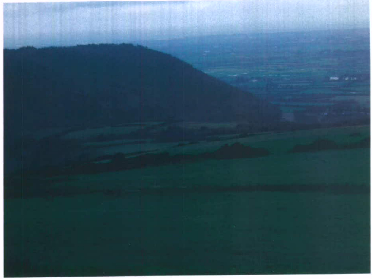 A low-resolution photograph depicting a rural landscape with a prominent hill on the left and fields extending into the distance.