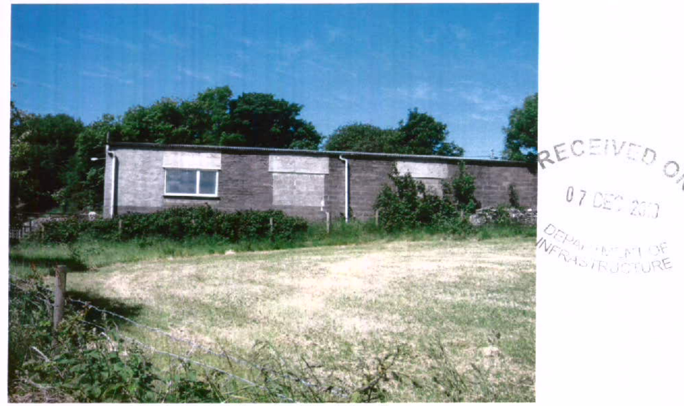 A photograph showing a long, single-story grey block building, likely a shed or outbuilding, situated in a grassy field with trees in the background.