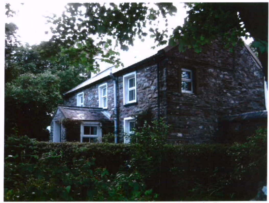 A photograph of a two-story stone house with white window frames, partially obscured by trees and a hedge.