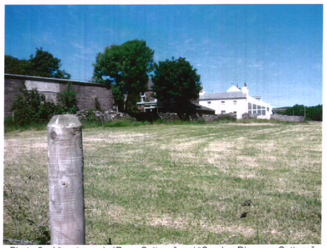 A photograph showing a grassy field in the foreground with a wooden post, looking towards a stone wall and white buildings in the background under a blue sky.