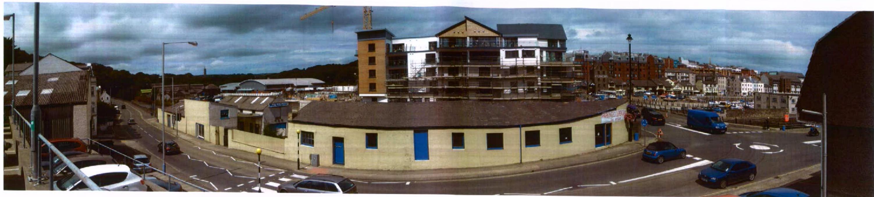A panoramic photograph showing a curved, single-story commercial building at a street junction, with a larger multi-story building under construction with scaffolding in the background.