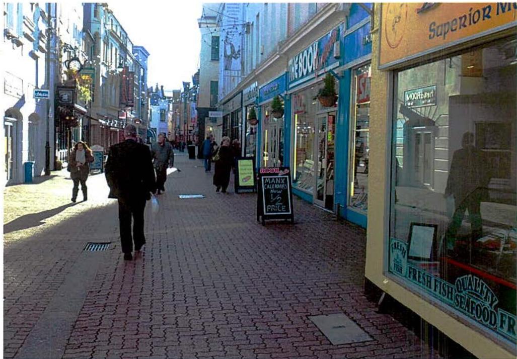 A street-level photograph of a pedestrianized high street in Douglas featuring brick paving, shop fronts, and A-frame advertising boards.
