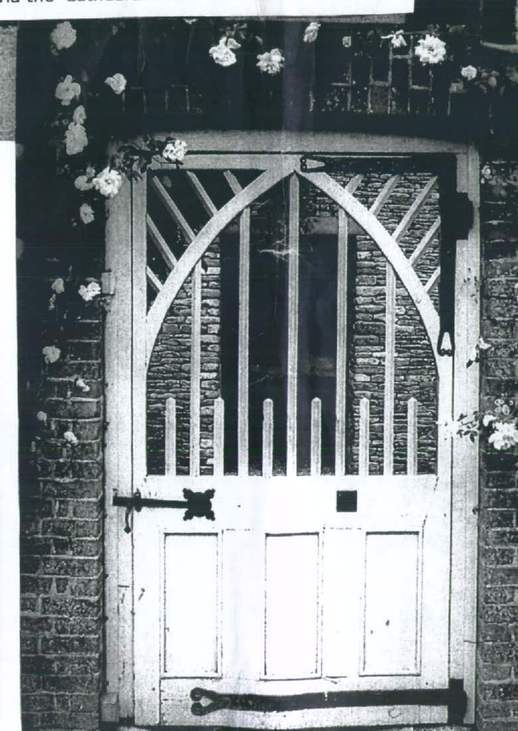 A black and white photograph showing a white wooden gate with an arched top and vertical slats, set within a stone wall surrounded by climbing flowers.