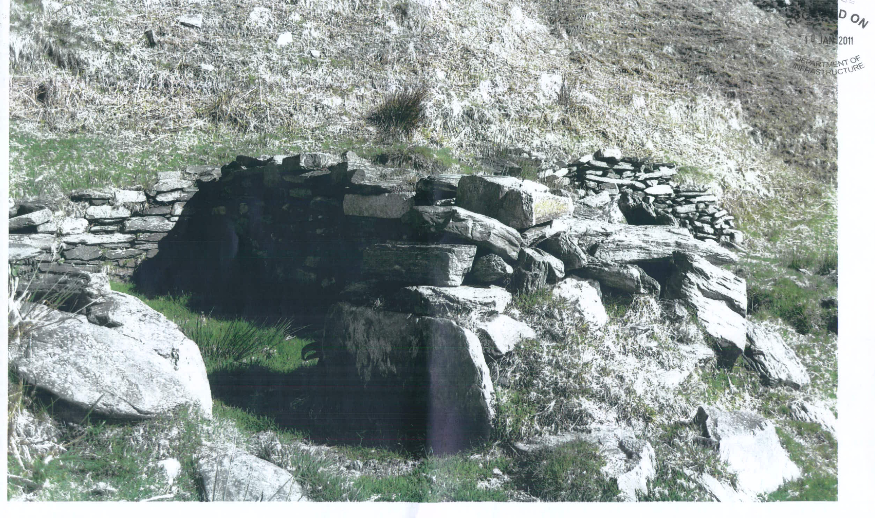 A photograph showing a small, dark stone structure built into a grassy, rocky hillside, consistent with the application description of a hermit's cell or keeill.