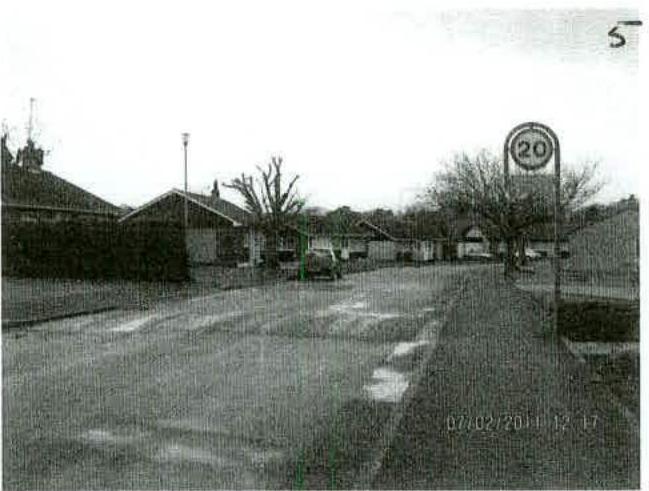 A grainy black and white photograph showing a street scene with a 20 mph speed limit sign, trees, and surrounding buildings.