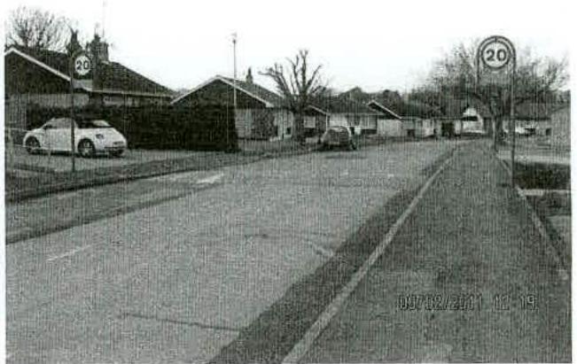 A grainy black and white photograph showing a street scene with single-story commercial buildings, parked cars, and 20 mph speed limit signs.