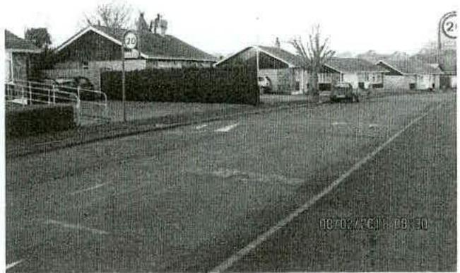Grainy black and white photograph showing a street scene with single-story buildings, parked cars, and a 20mph speed limit sign.
